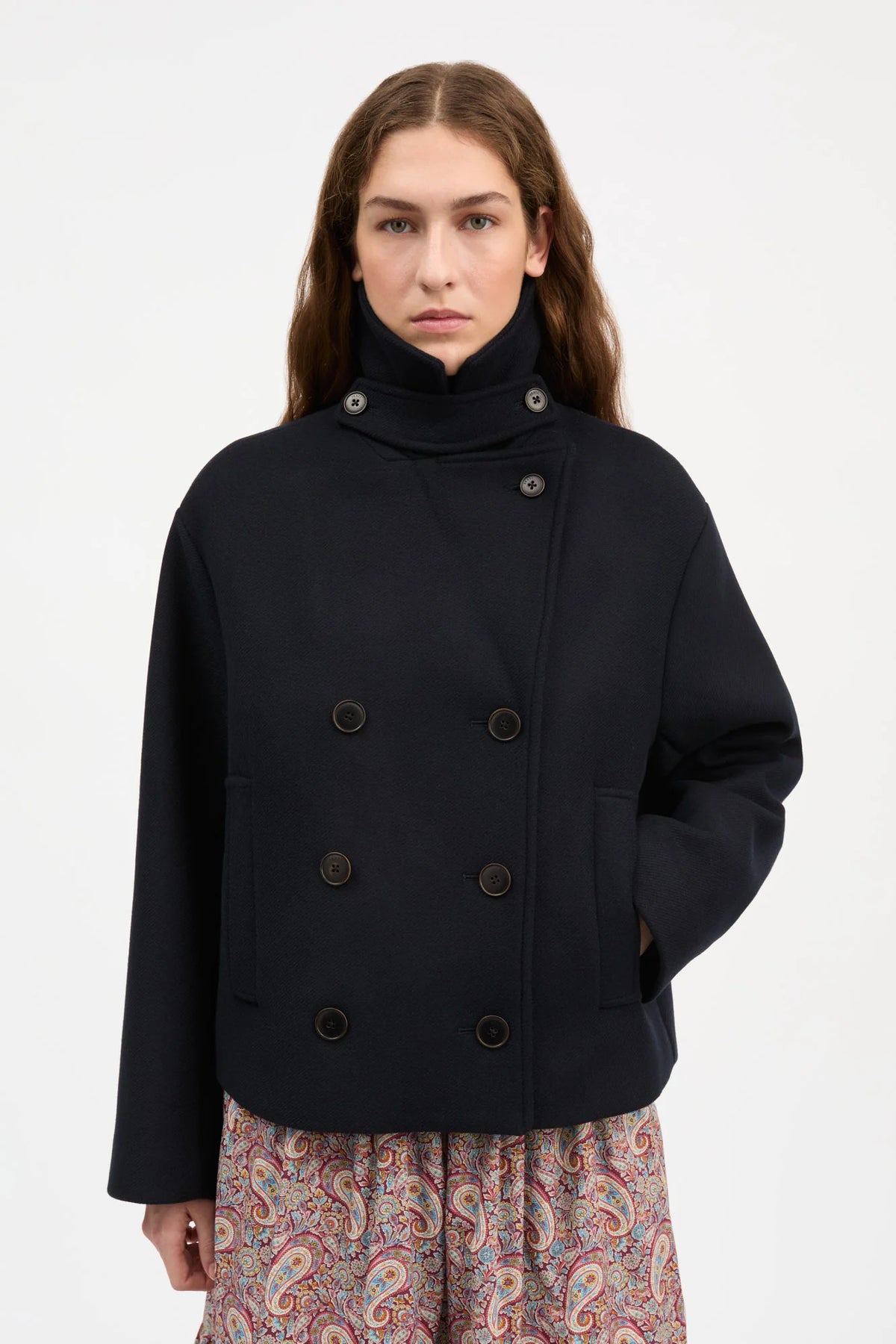 A woman modeling a dark navy blue double-breasted coat with a high mock neck and dark buttons, paired with a paisley skirt, against a plain light gray background.