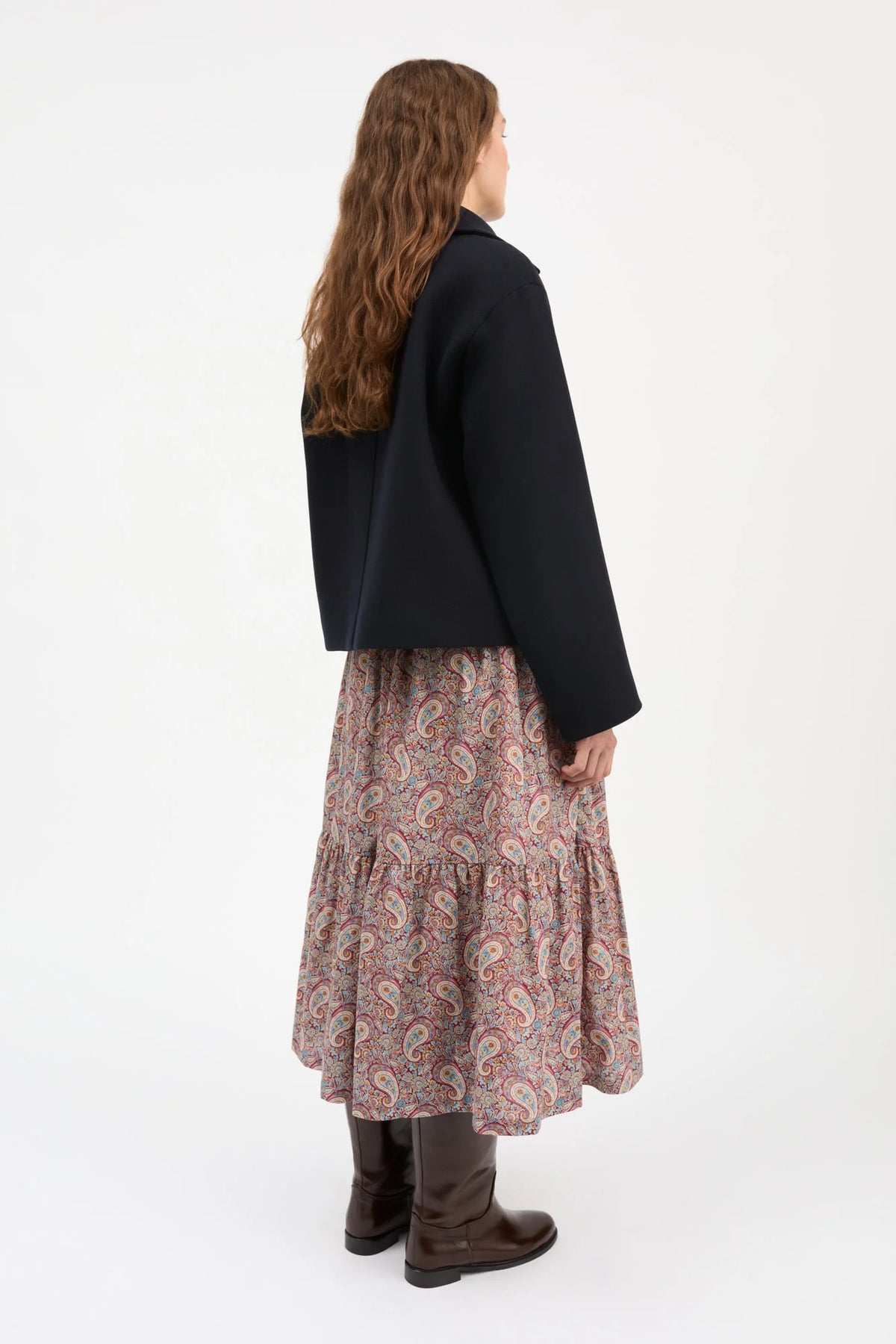 A woman is shown from the back wearing a black cropped jacket, a long tiered skirt with a red, blue, and beige paisley pattern, and dark brown boots, set against a plain white studio background.
