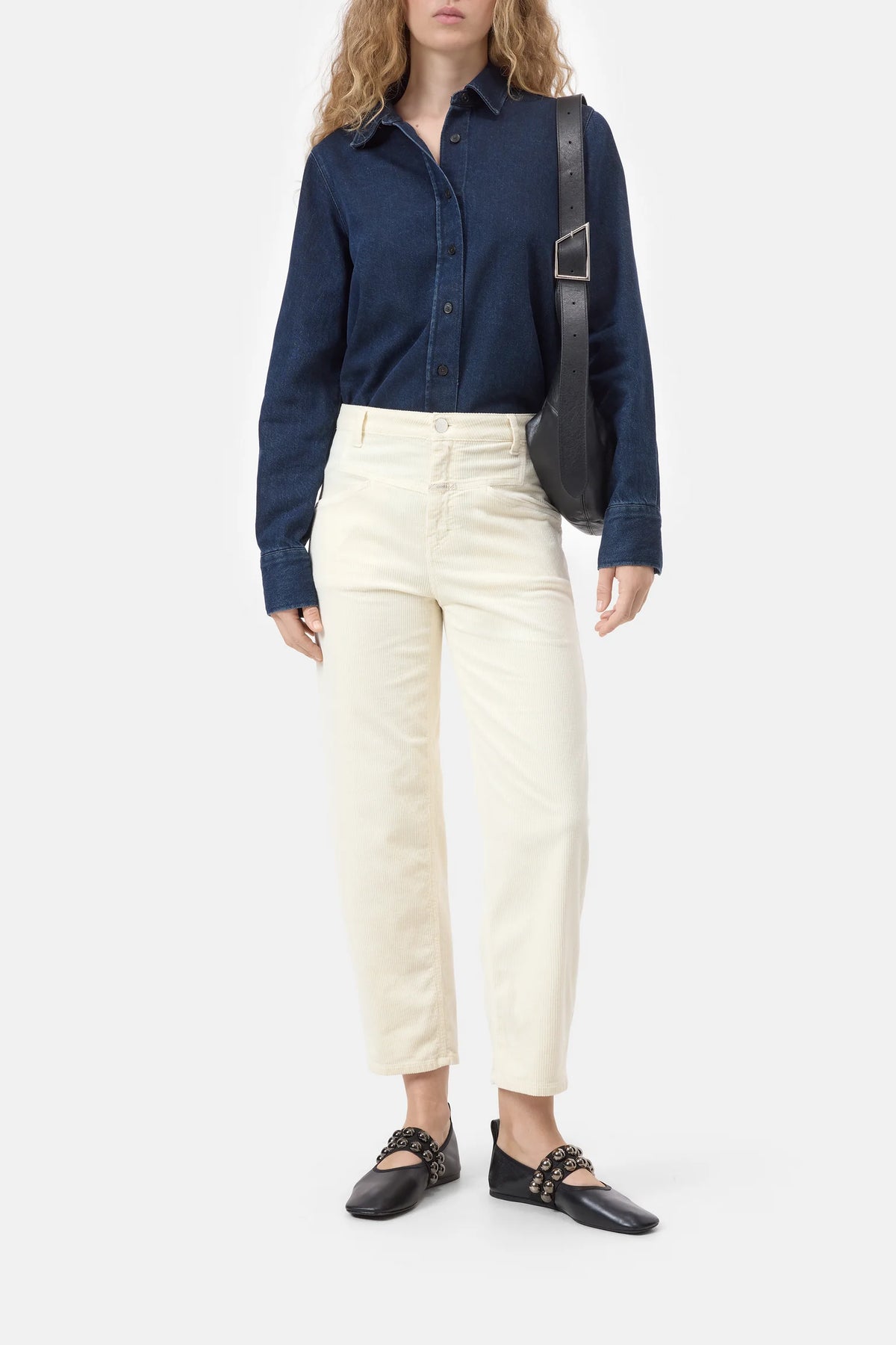 A model wearing a dark blue denim button-up shirt, off-white cropped trousers, and black leather flats with gold chain details, carrying a black shoulder bag against a light gray background.