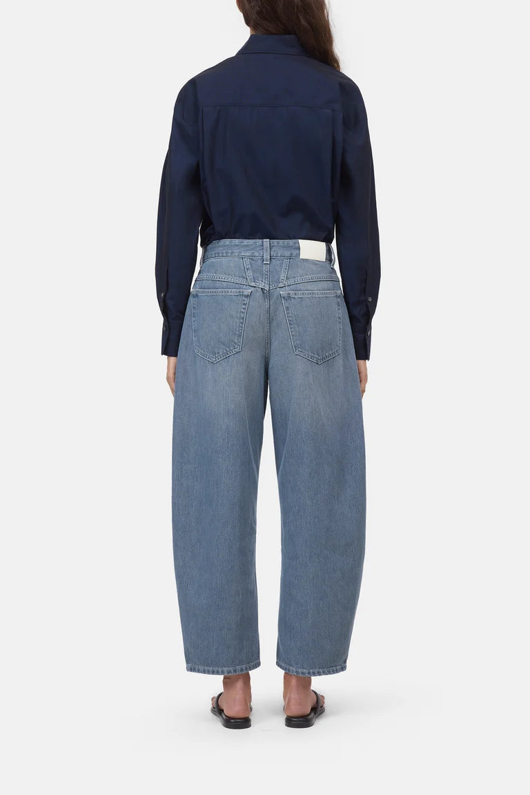 Rear view of a model wearing a dark navy long-sleeve button-up shirt and light blue wide-leg cropped jeans, paired with black flat sandals, against a light gray studio background.