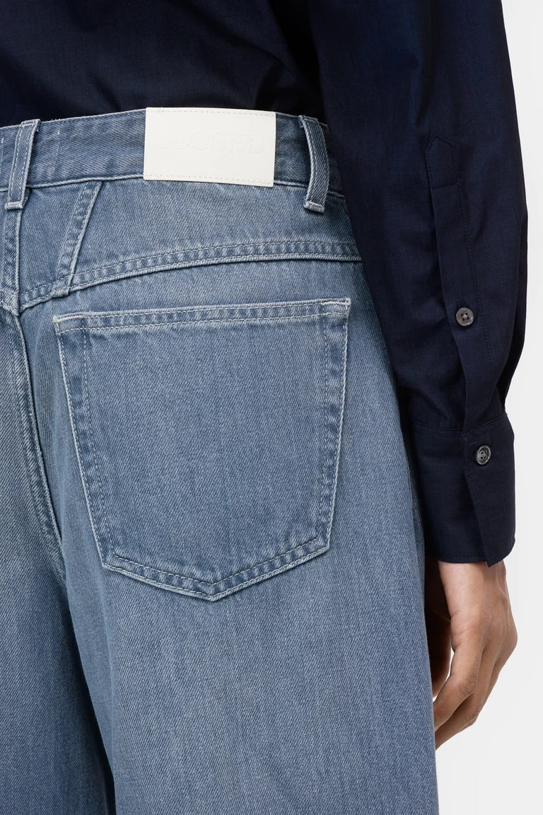 Close-up of the back waistband and pocket area of light blue denim jeans, worn with a dark navy button-up shirt against a plain light background.