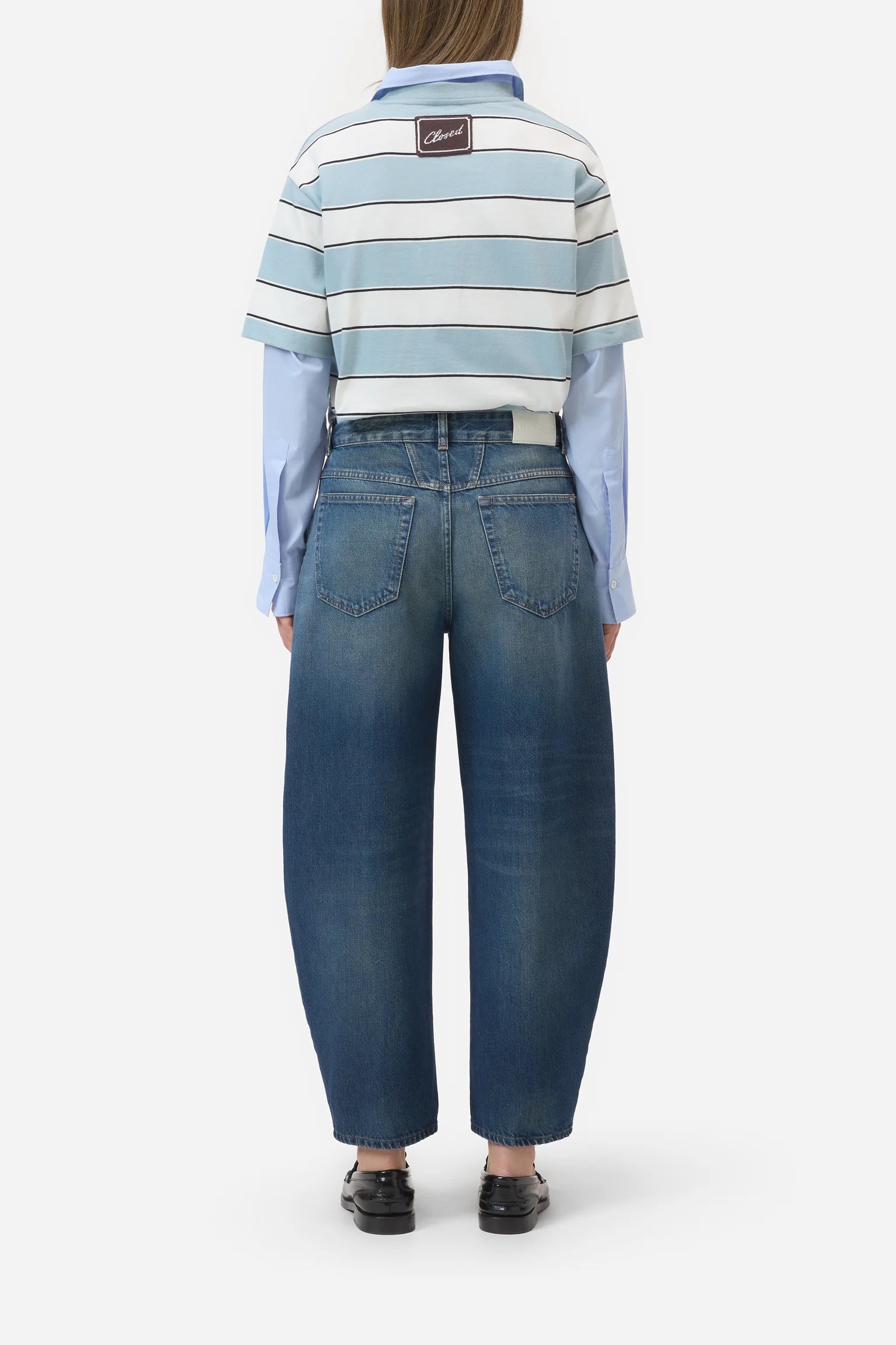 A model viewed from the back wearing a light blue and white striped short-sleeve polo shirt layered over a light blue long-sleeve shirt, paired with wide-leg blue denim jeans and black shoes, against a plain light gray background.