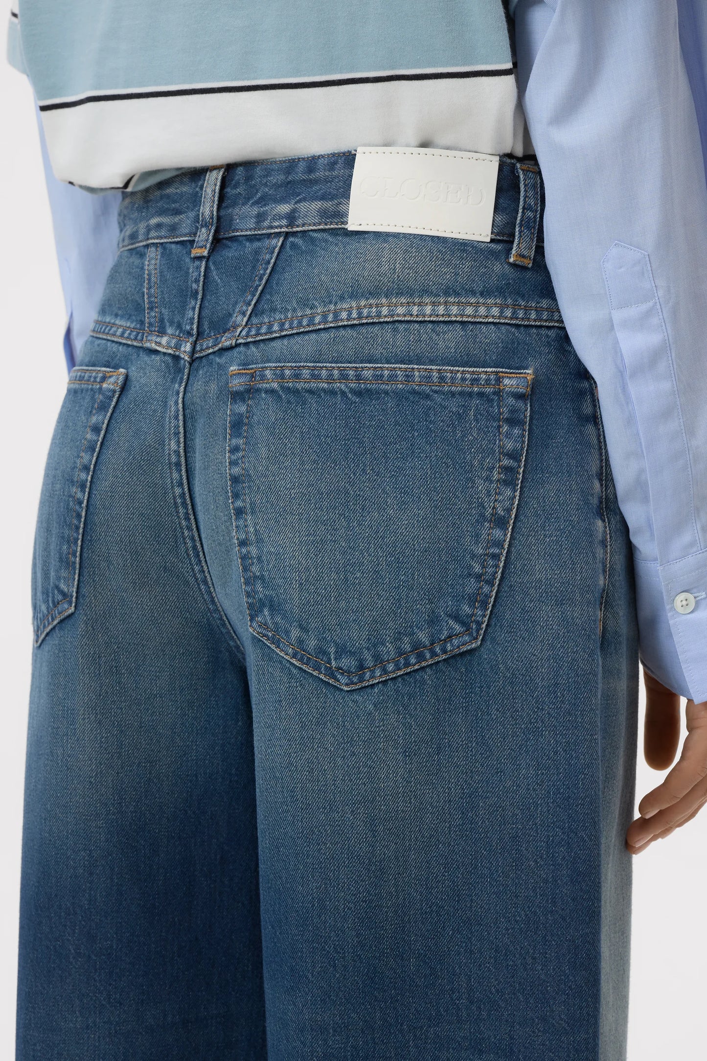 Close-up back view of a person wearing blue denim jeans with a light blue shirt tucked in, against a plain light background.