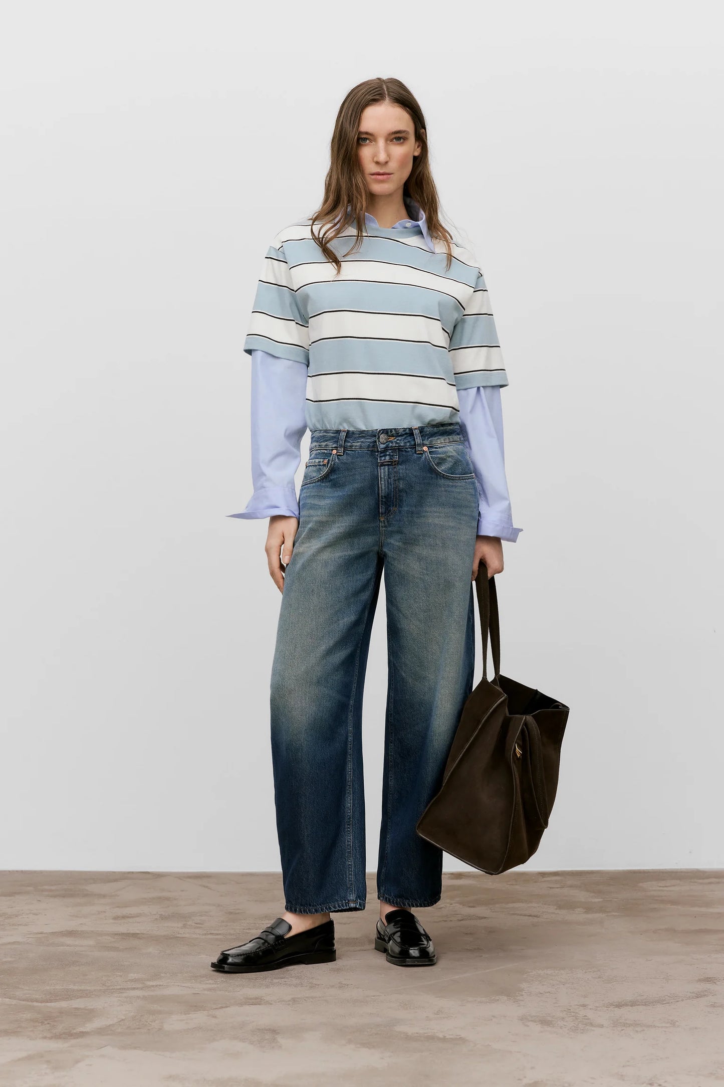 Woman wearing a striped sweater and jeans, holding a brown bag on a plain background