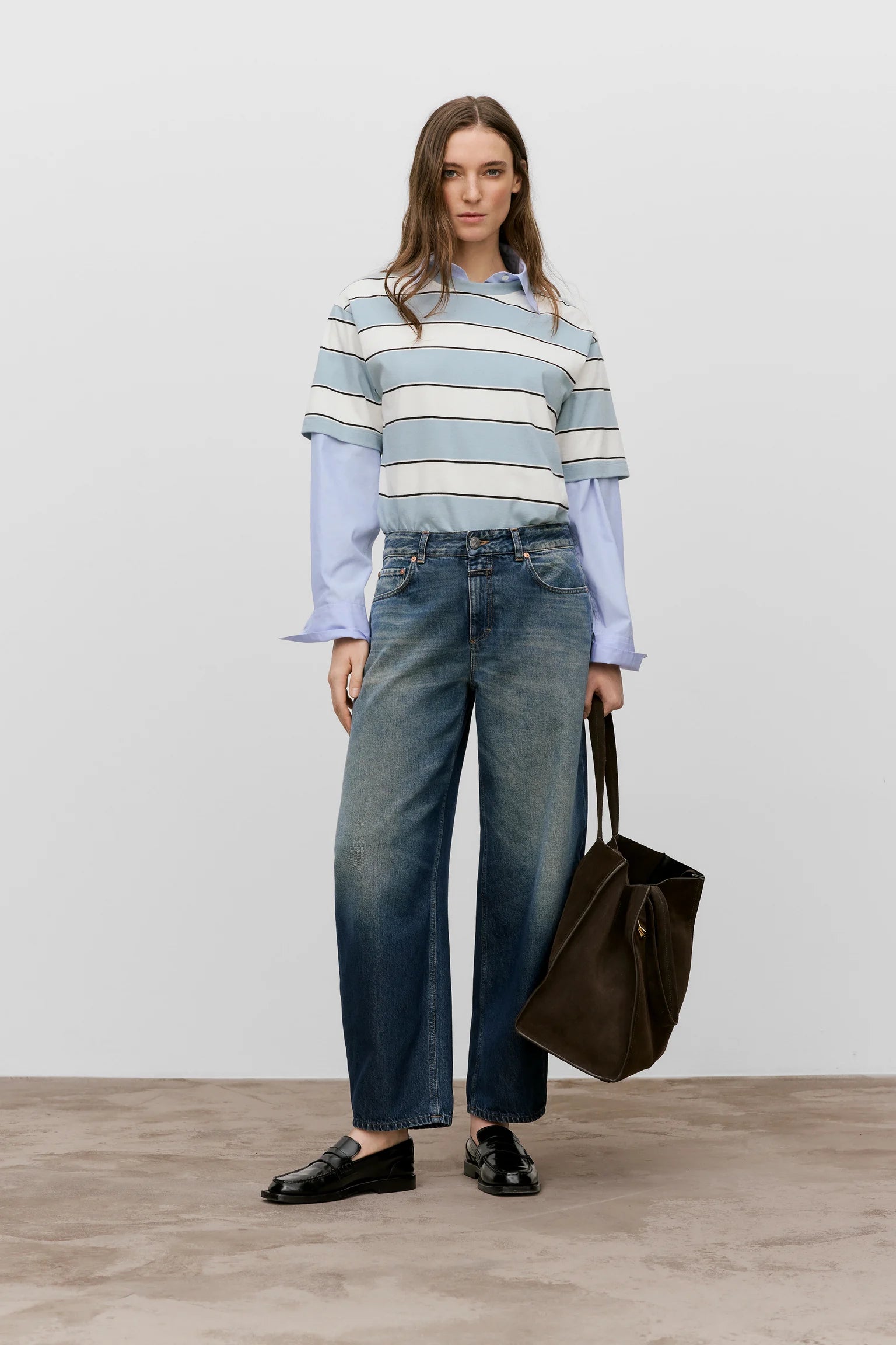Woman wearing a striped sweater and jeans, holding a brown bag on a plain background
