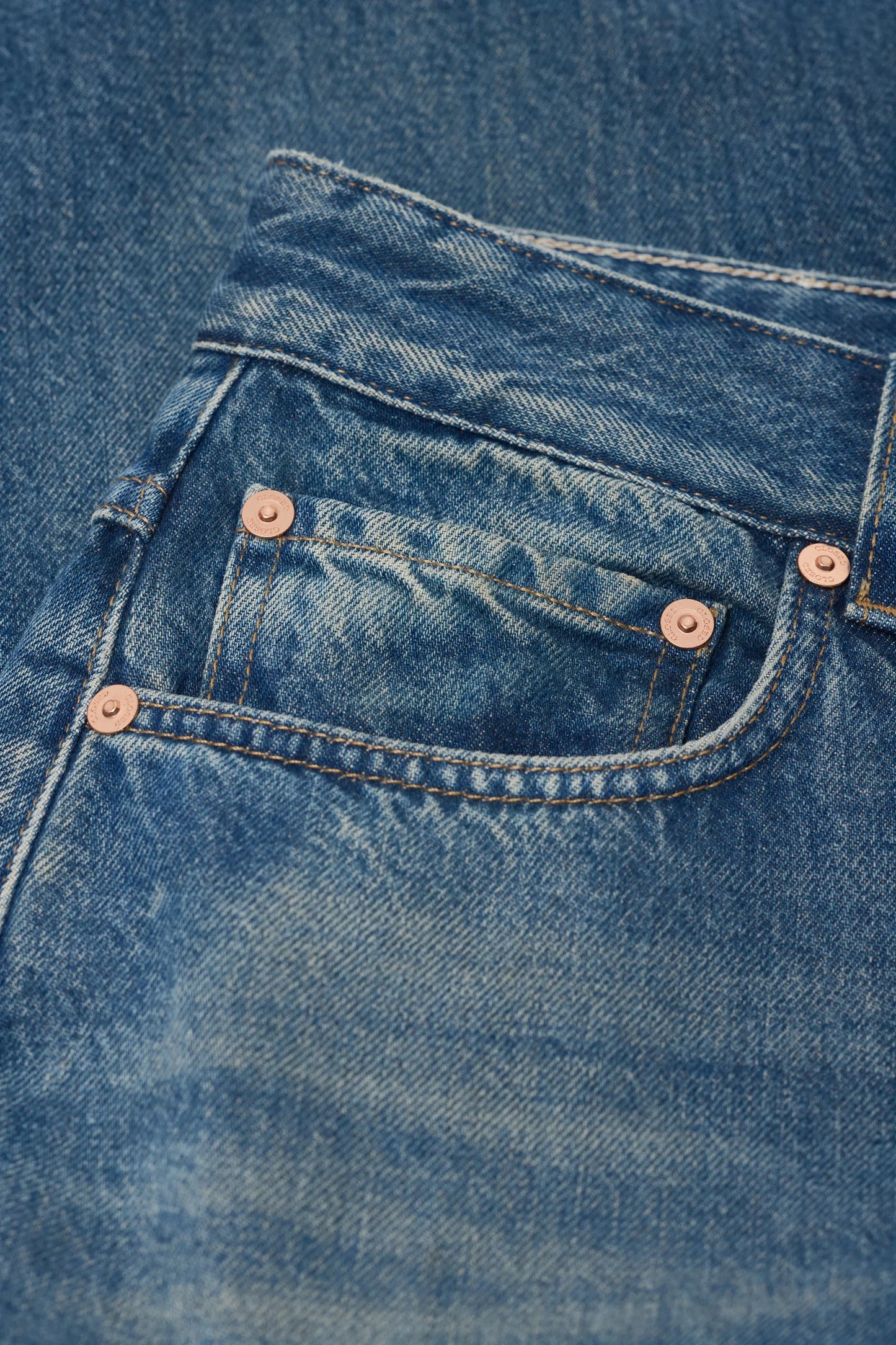Close-up of a blue denim pair of jeans showing the back pocket with copper-toned rivets and stitching.
