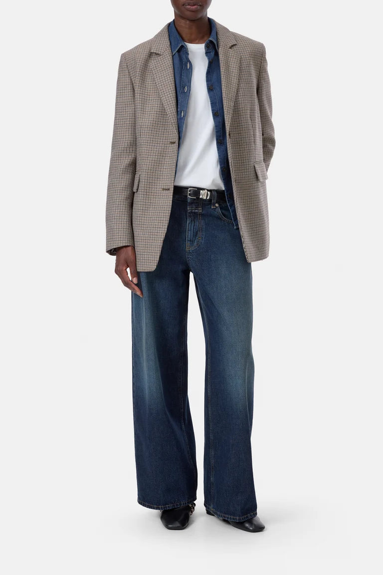 A model wearing a beige and brown checkered blazer, a white t-shirt, a blue denim shirt, wide-leg blue jeans, and black shoes, photographed against a light gray studio background.