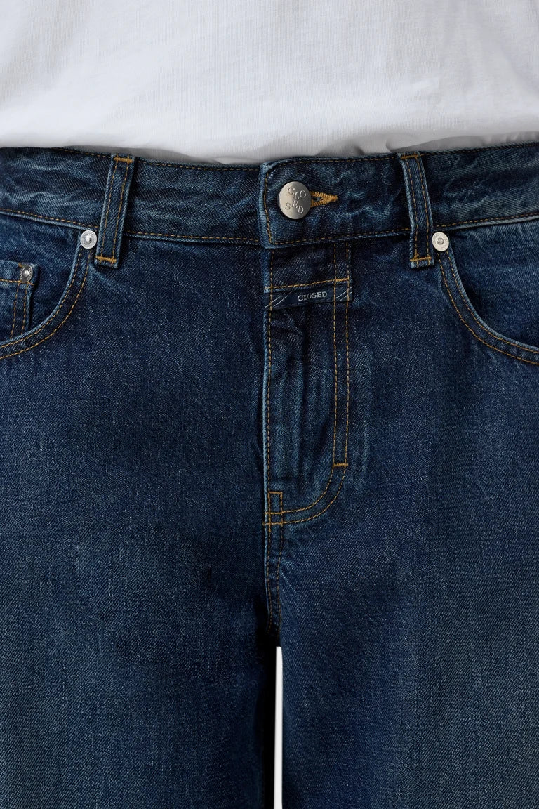 Close-up of dark blue denim jeans worn with a white top, showing the waistband, button, belt loops, front pockets, and stitching details.