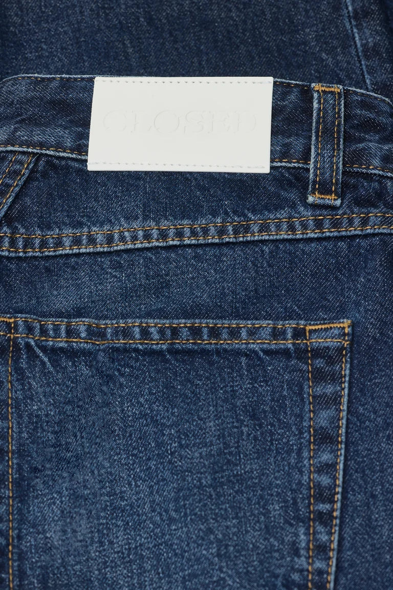 Close-up of the back waistband of dark blue denim jeans, showing a white rectangular brand patch with debossed lettering and contrast orange stitching.