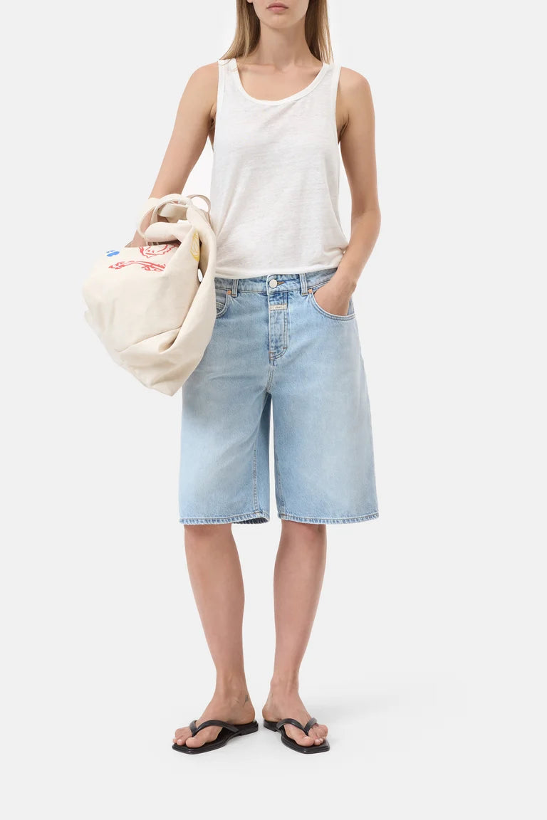Person wearing a white tank top and light blue denim shorts, holding a beige bag on a white background