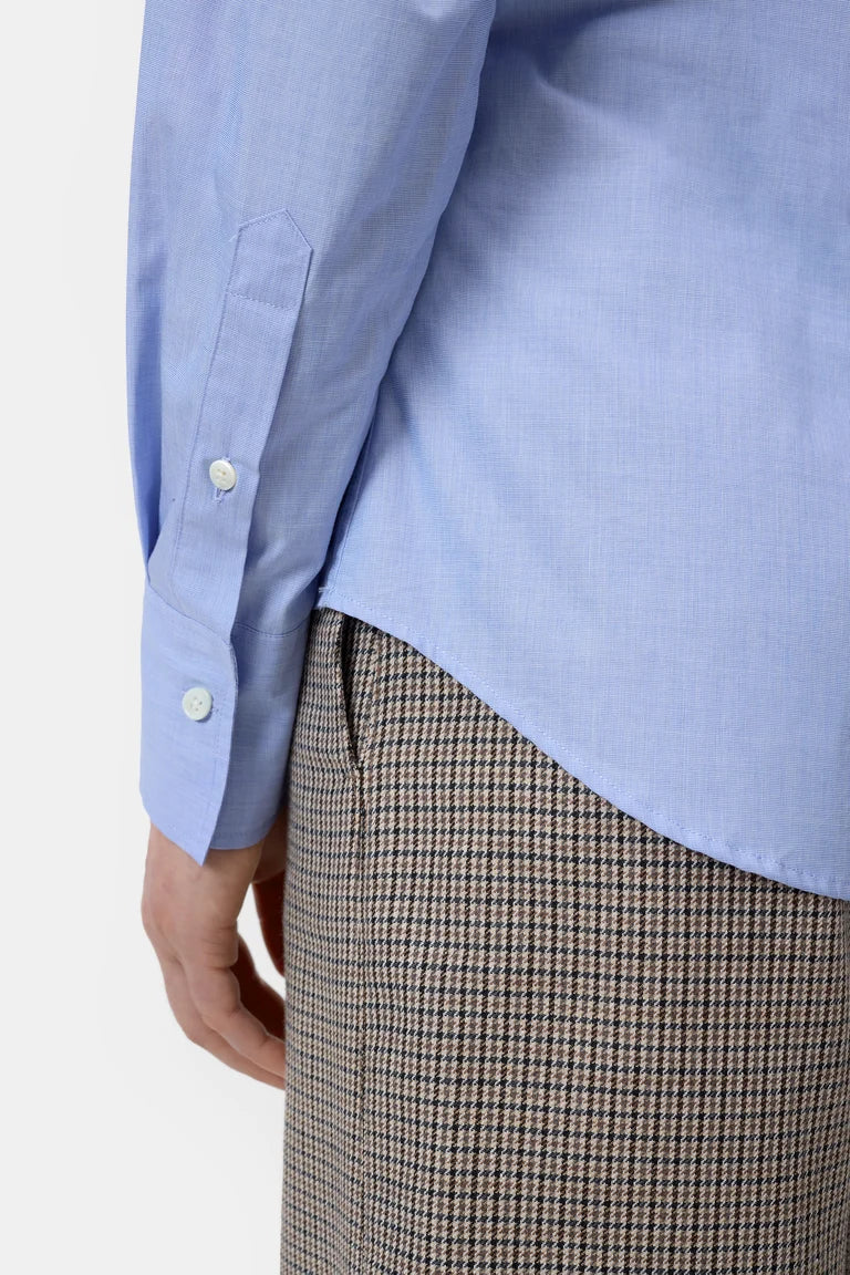 Close-up of a person wearing a light blue long-sleeve button-up shirt and brown-and-white houndstooth trousers, cropped to show the shirt cuff, hand, and lower torso against a plain light background.