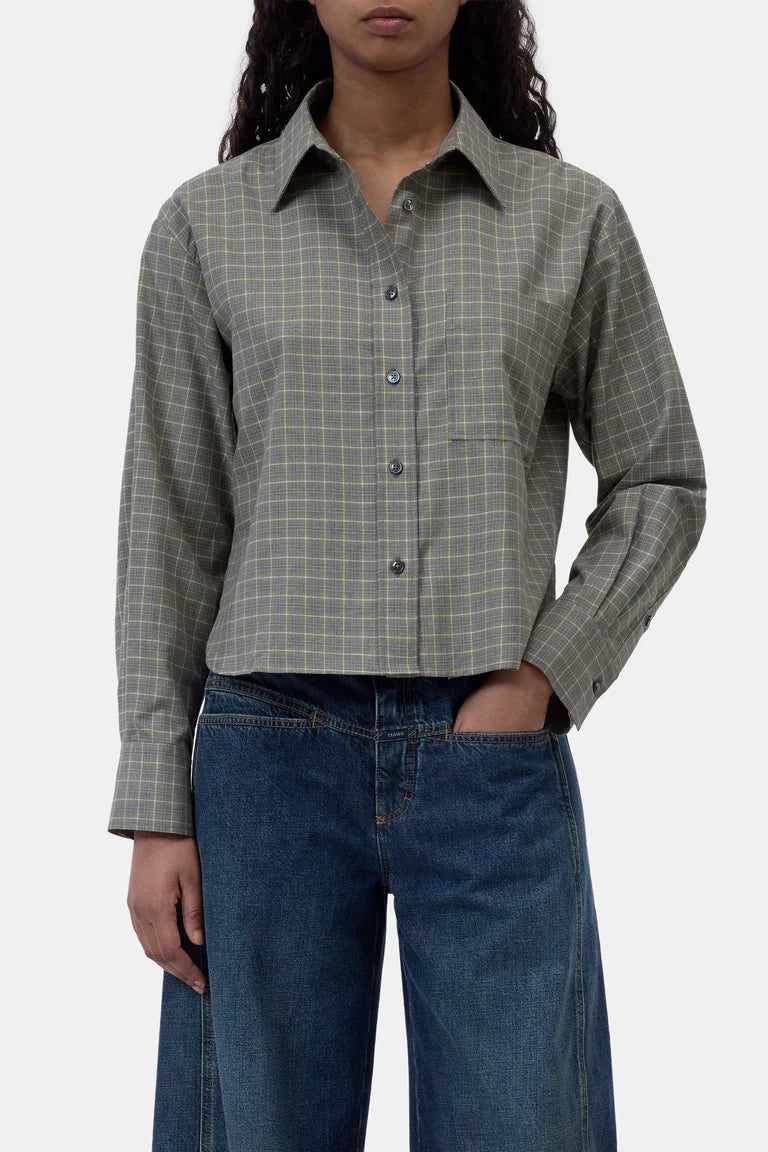 A woman wearing a cropped, long-sleeve button-up shirt with a muted green and gray windowpane plaid pattern, paired with blue jeans, against a plain light gray background.