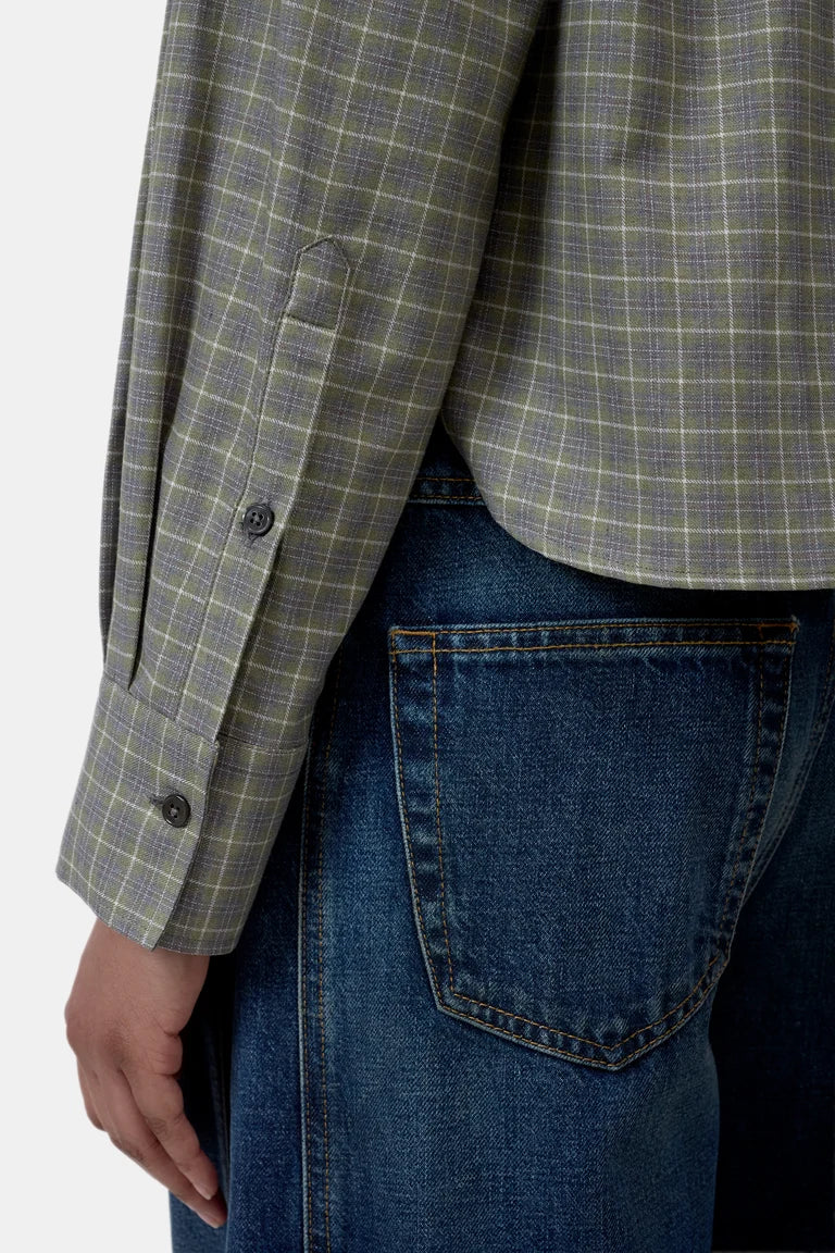 Close-up of a person wearing a muted green and gray plaid button-up shirt with dark blue jeans, showing the shirt cuff, buttons, and the back pocket of the jeans.