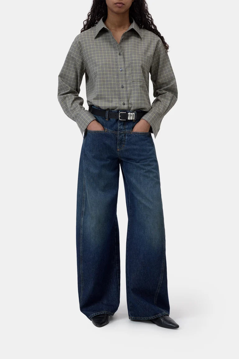 A model wearing a gray and green plaid button-down shirt tucked into dark blue wide-leg denim jeans with a black belt, photographed against a plain light gray studio background.