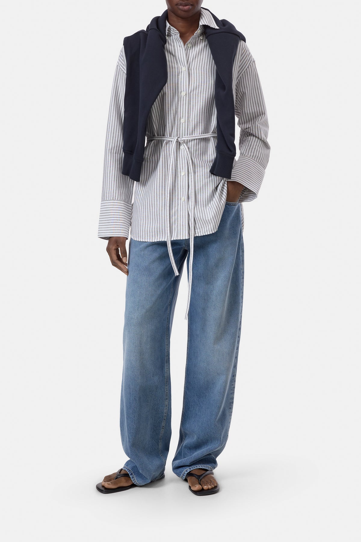 A model wearing a styled outfit consisting of a white and navy striped button-down shirt, a navy sleeveless vest, wide-leg blue jeans, and brown leather sandals, shot against a plain white studio background.