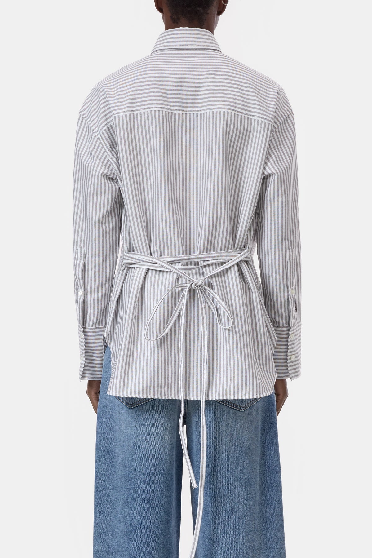A person shown from the back wearing a long-sleeve white shirt with thin gray vertical stripes and a matching self-tie belt, paired with wide-leg blue jeans against a plain light gray studio background.