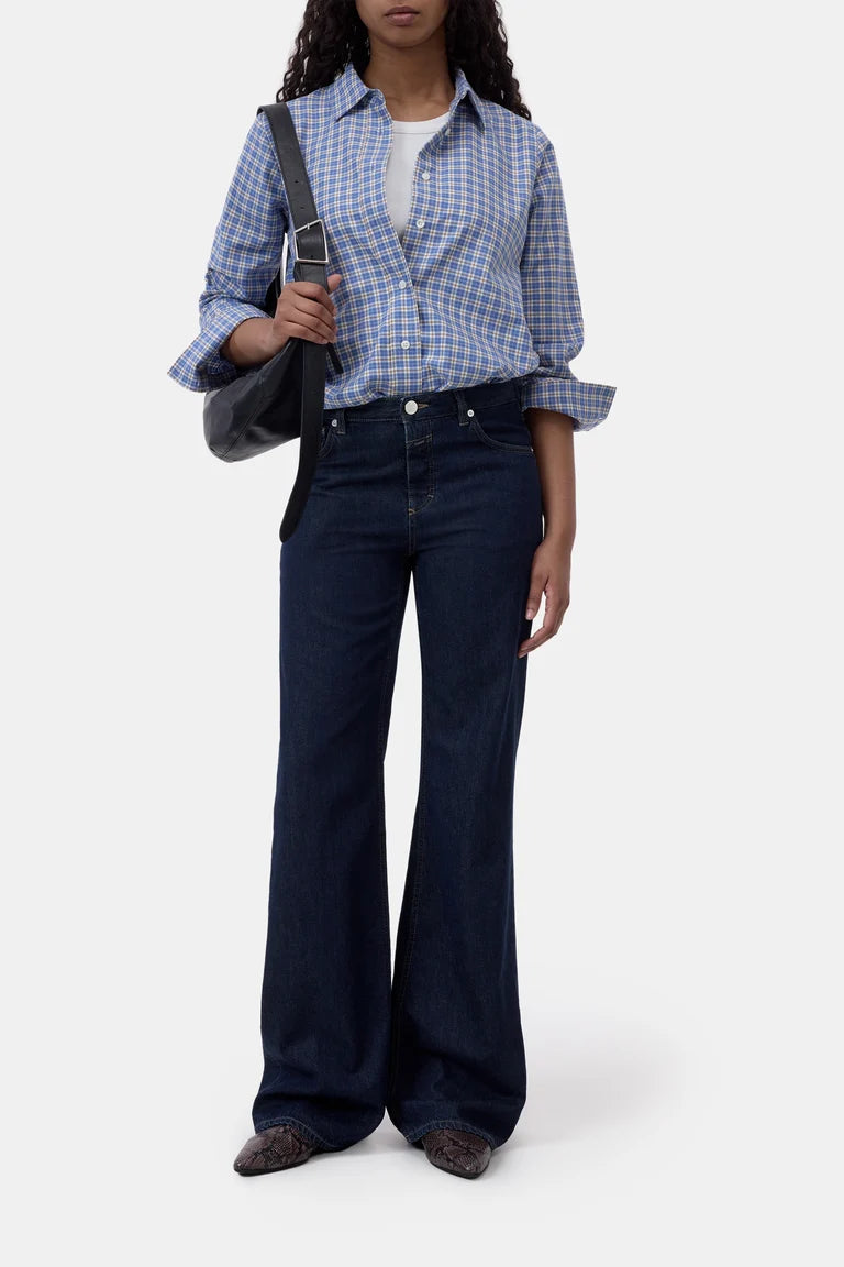 A model wearing a blue and white checkered button-down shirt, dark blue wide-leg jeans, and brown pointed-toe flats, carrying a black shoulder bag against a light gray background.