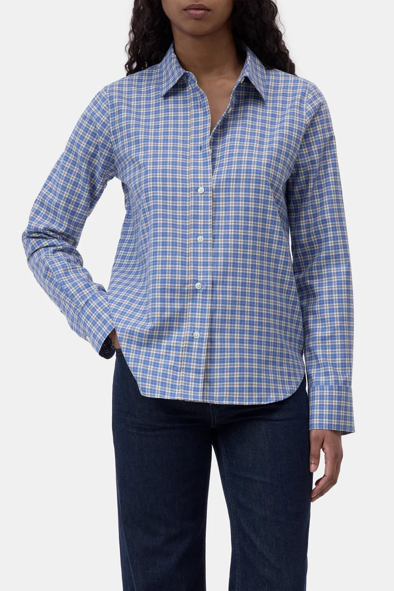 A woman wearing a blue and white checkered long-sleeve button-down shirt with dark blue jeans, shown from the neck down against a light gray background.