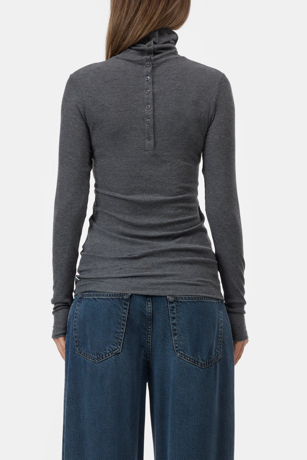 A woman shown from the back wearing a dark gray long-sleeve turtleneck top with a buttoned collar detail and blue jeans, against a light gray studio background.