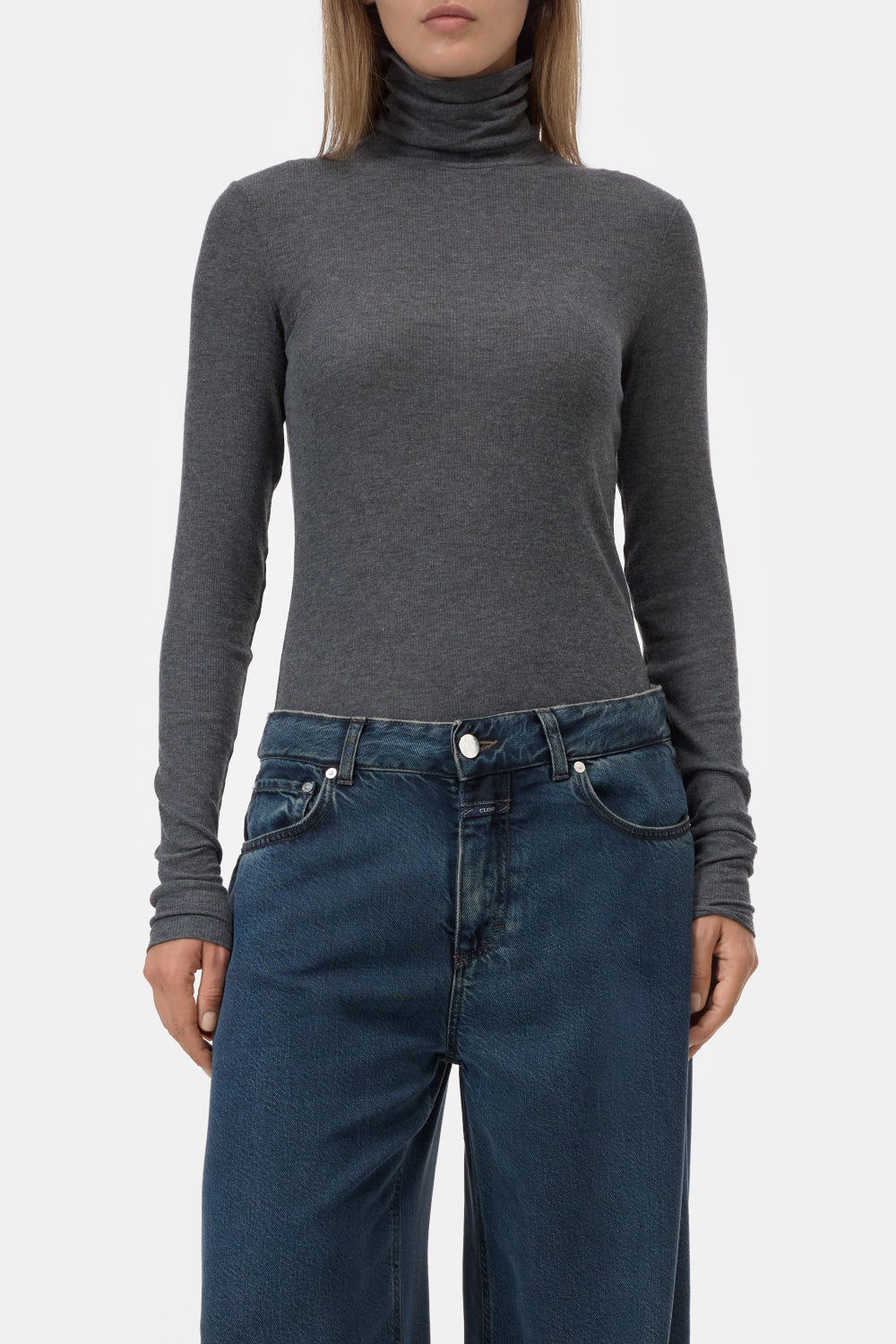 A woman wearing a dark gray long-sleeve turtleneck top and dark blue jeans, shown from the neck down against a light gray studio background.