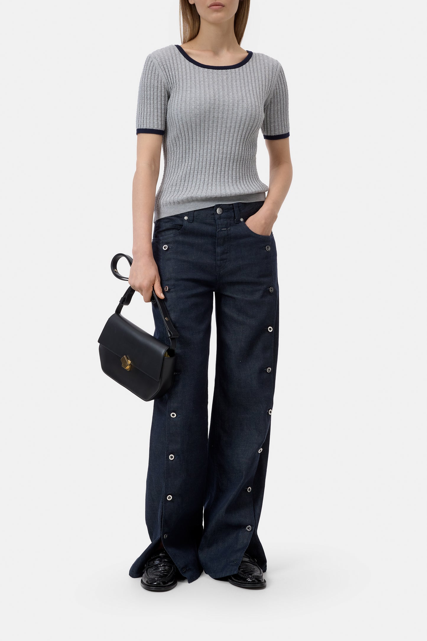Person wearing a gray ribbed sweater and dark blue jeans holding a black handbag on a white background