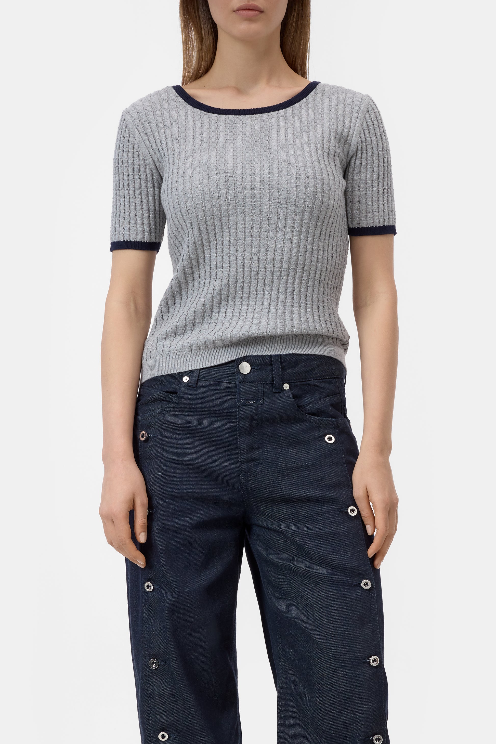 Woman wearing a gray ribbed sweater and dark blue pants with button details on a white background
