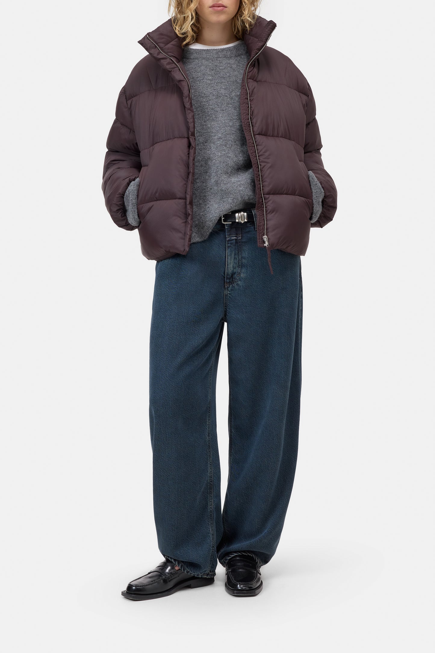 A model wearing a dark plum-brown puffer jacket over a gray sweater, paired with wide-leg dark blue jeans and black leather shoes, photographed against a light gray studio background.