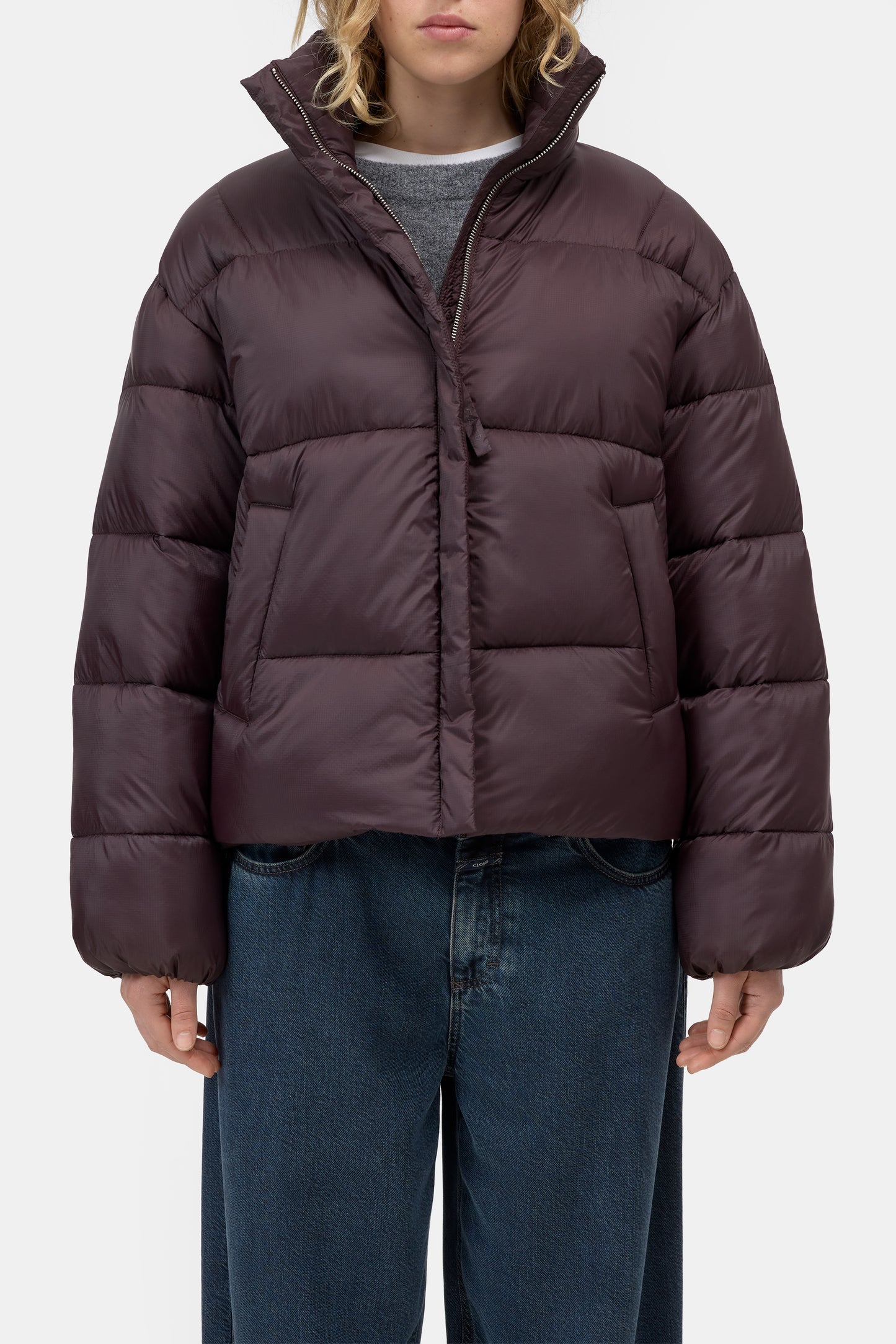 A model wearing a dark plum purple cropped puffer jacket with a high stand-up collar, paired with blue jeans and a gray t-shirt, against a plain light gray background.