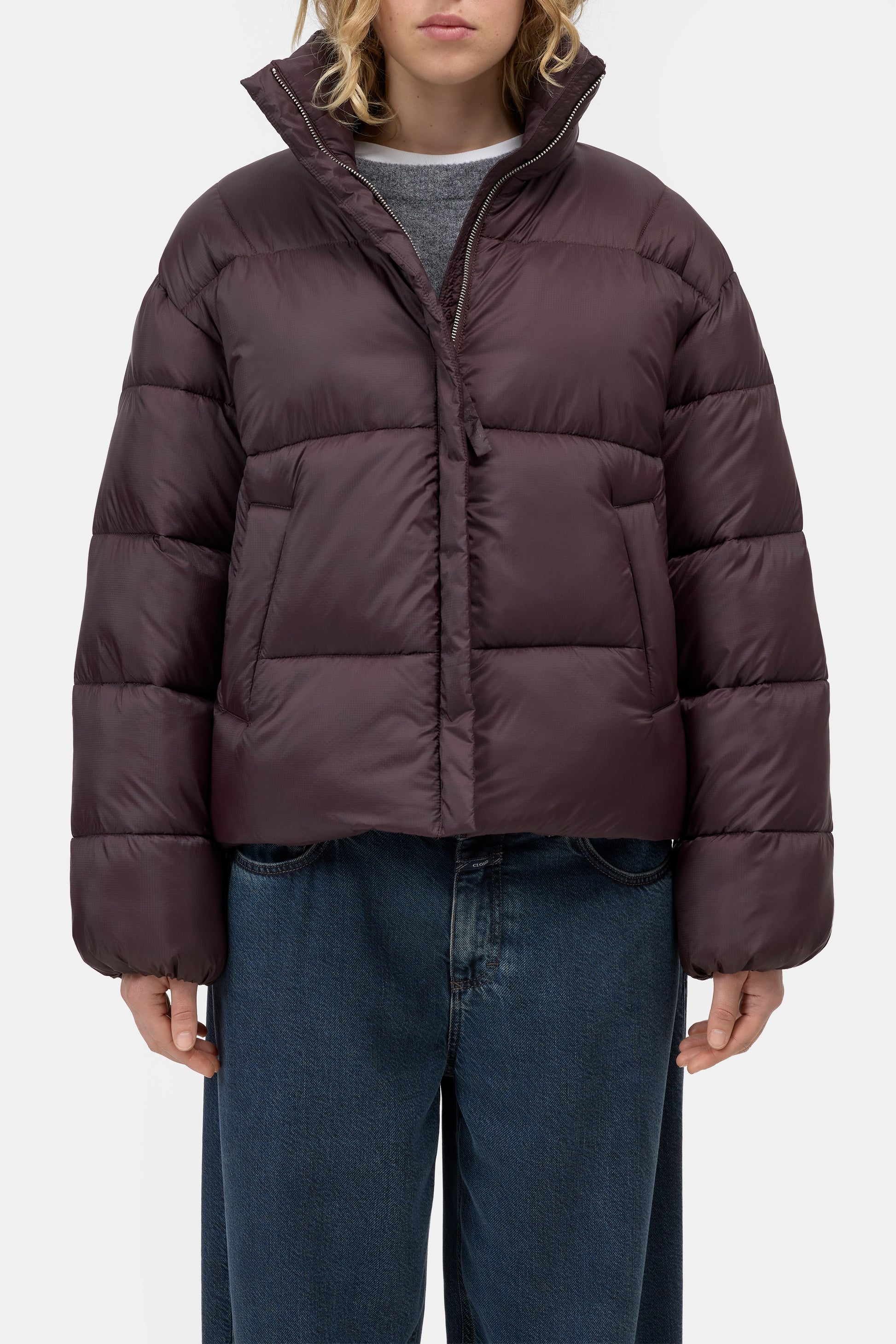 A model wearing a dark plum purple cropped puffer jacket with a high stand-up collar, paired with blue jeans and a gray t-shirt, against a plain light gray background.
