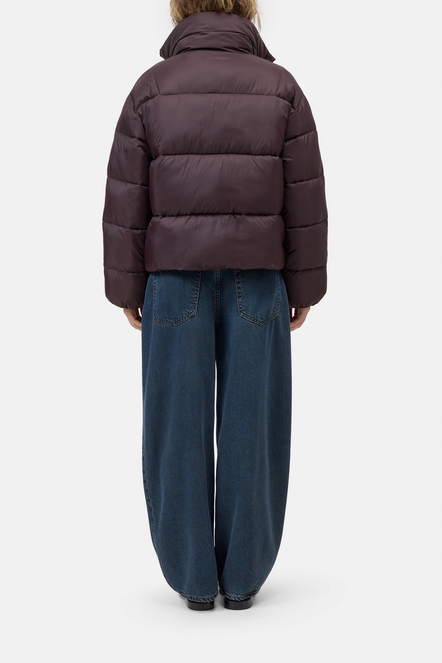A model shown from the back wearing a dark brown cropped puffer jacket and wide-leg blue jeans against a light gray studio background.