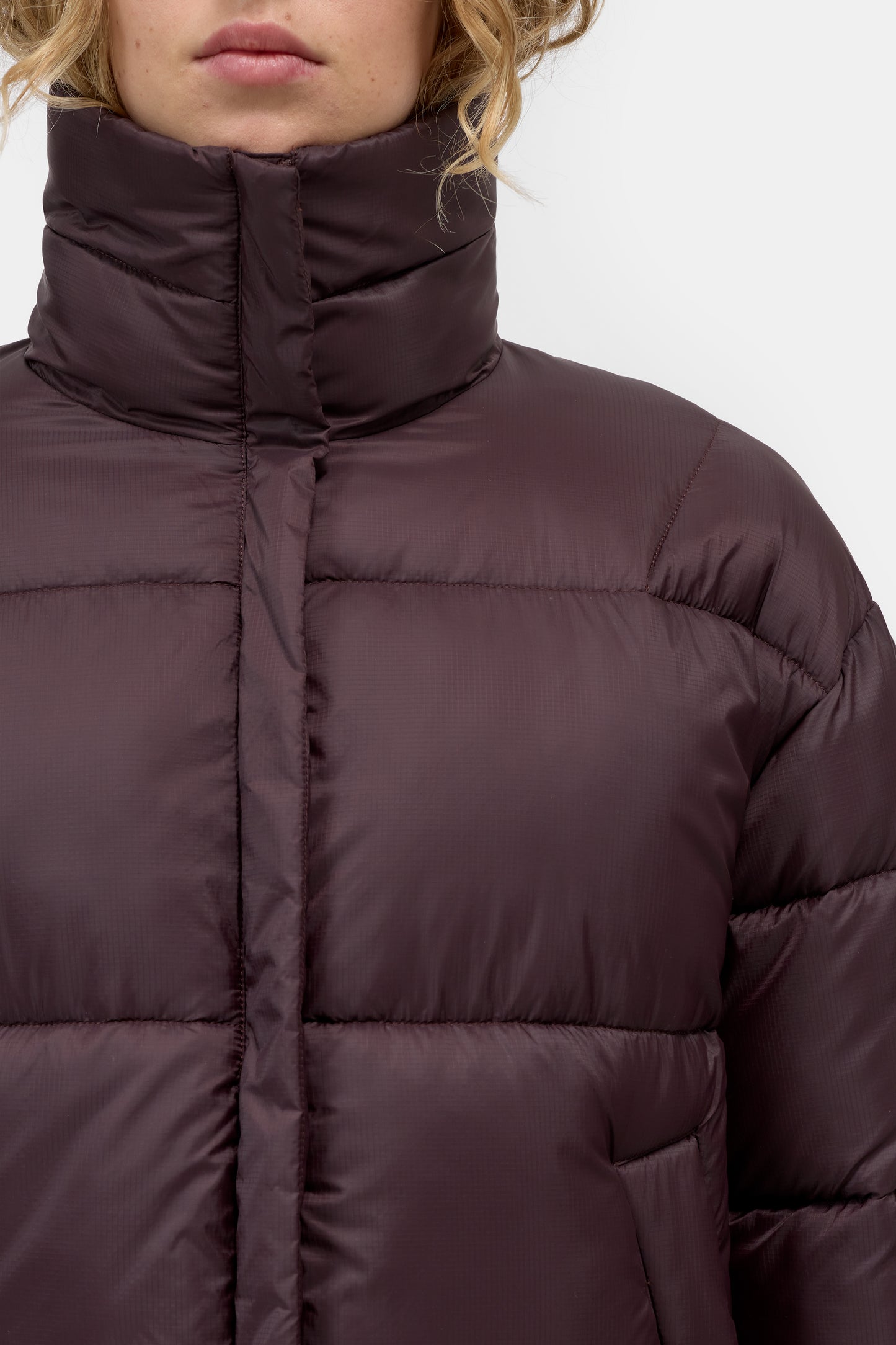 Close-up of a model wearing a dark plum purple puffer jacket with a high collar and horizontal quilted stitching.