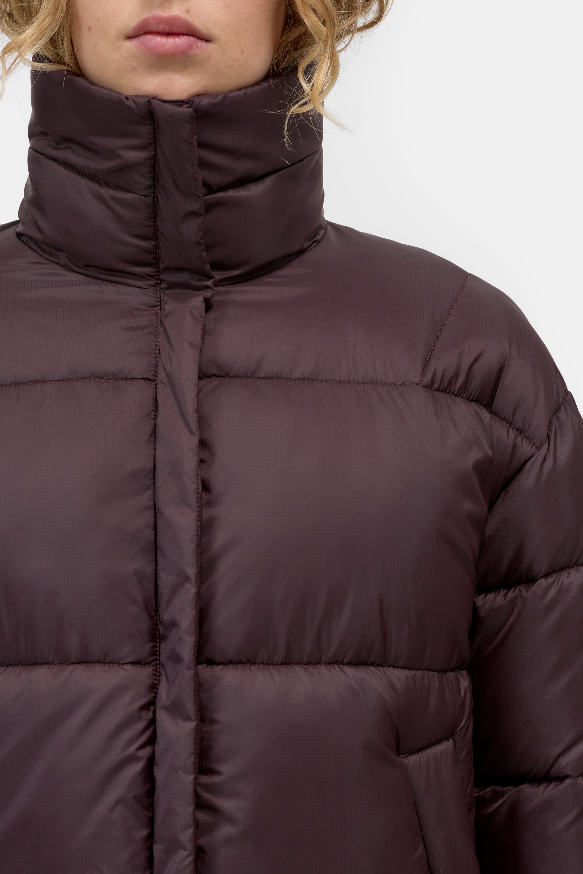 Close-up of a model wearing a dark plum purple puffer jacket with a high collar and horizontal quilted stitching.