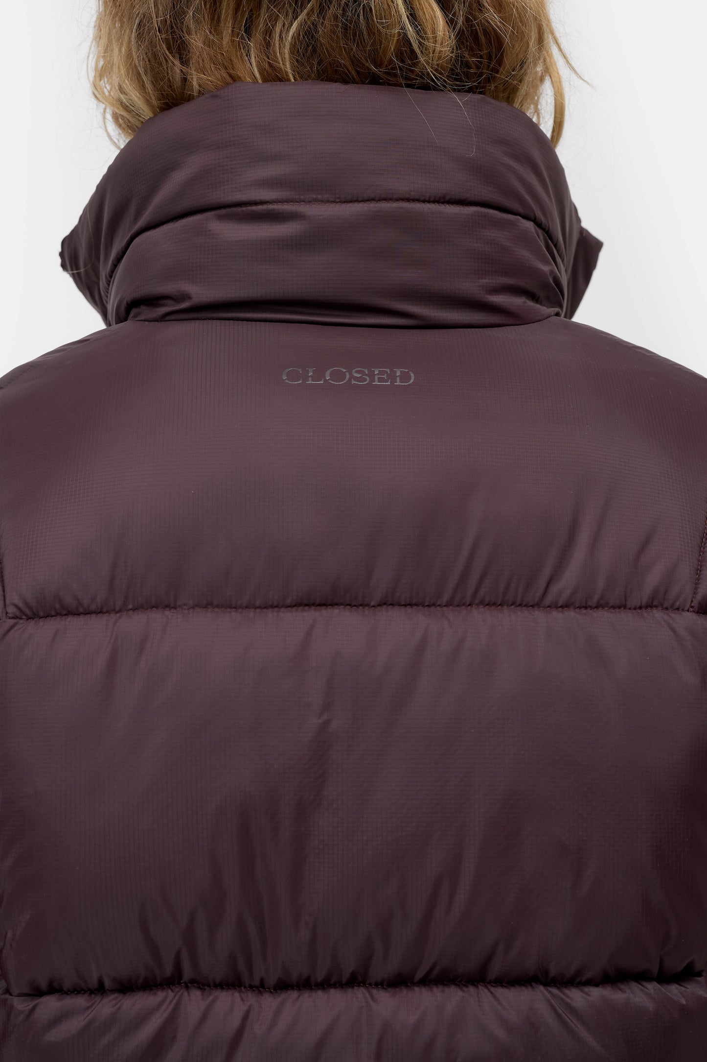 Close-up back view of a person wearing a dark plum puffer jacket with a high padded collar and the word 'CLOSED' printed on the upper back.
