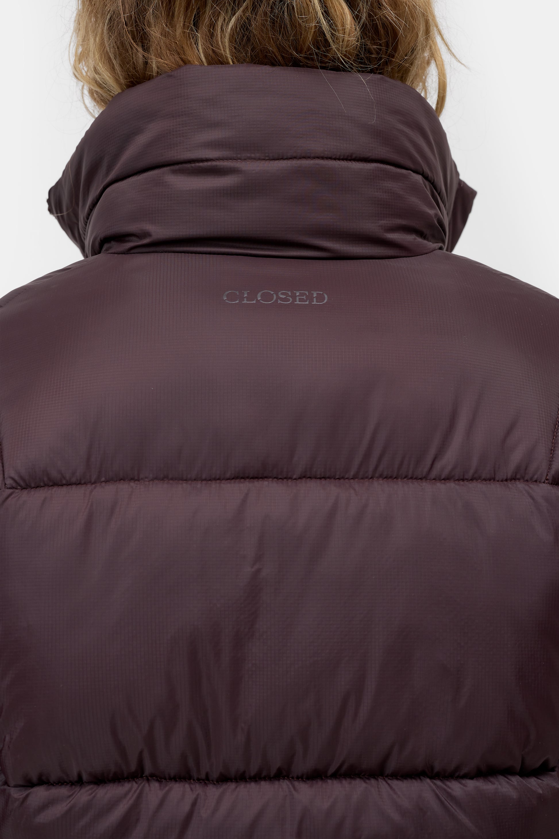 Close-up back view of a person wearing a dark plum puffer jacket with a high padded collar and the word 'CLOSED' printed on the upper back.