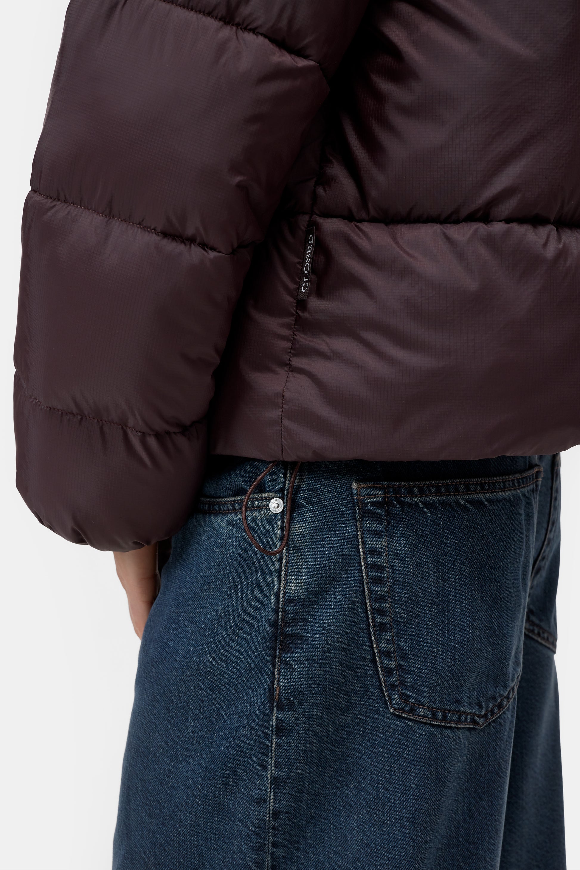 Close-up of a person wearing a dark brown puffer jacket and blue denim jeans, showing the back pocket and waist area against a light gray background.