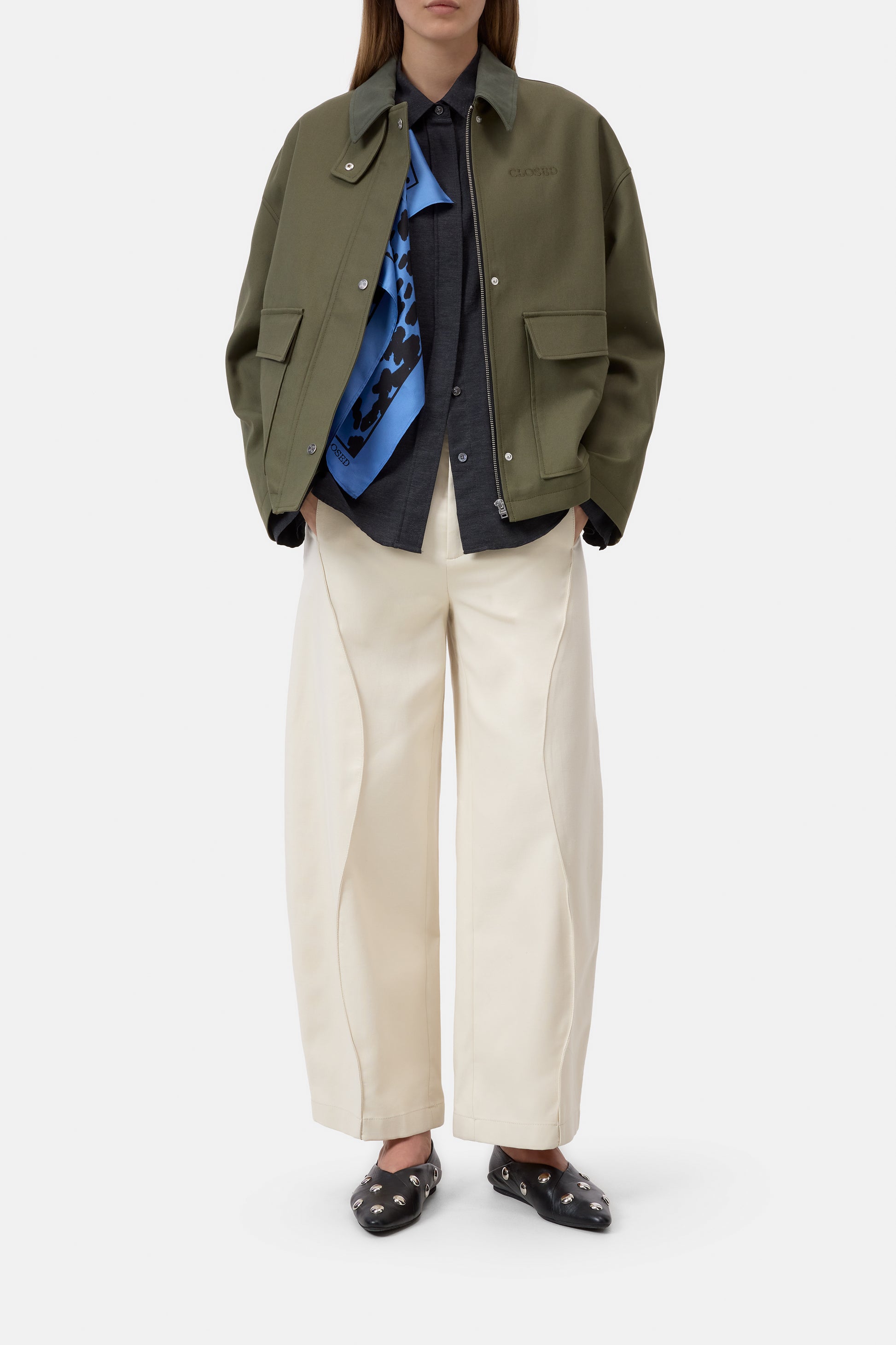 Person wearing an olive green jacket, blue shirt, black sweater, beige pants, and black shoes on a white background
