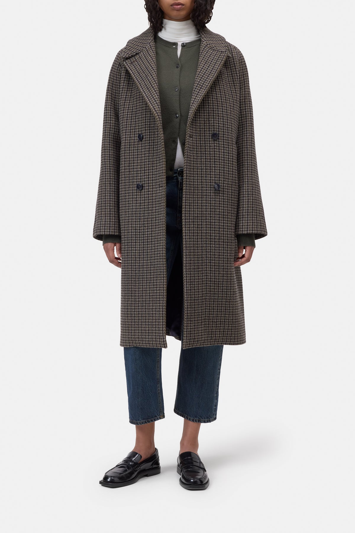 A model wearing a long double-breasted houndstooth coat in brown and beige, styled with a green cardigan, white turtleneck, blue jeans, and black loafers, against a light gray background.