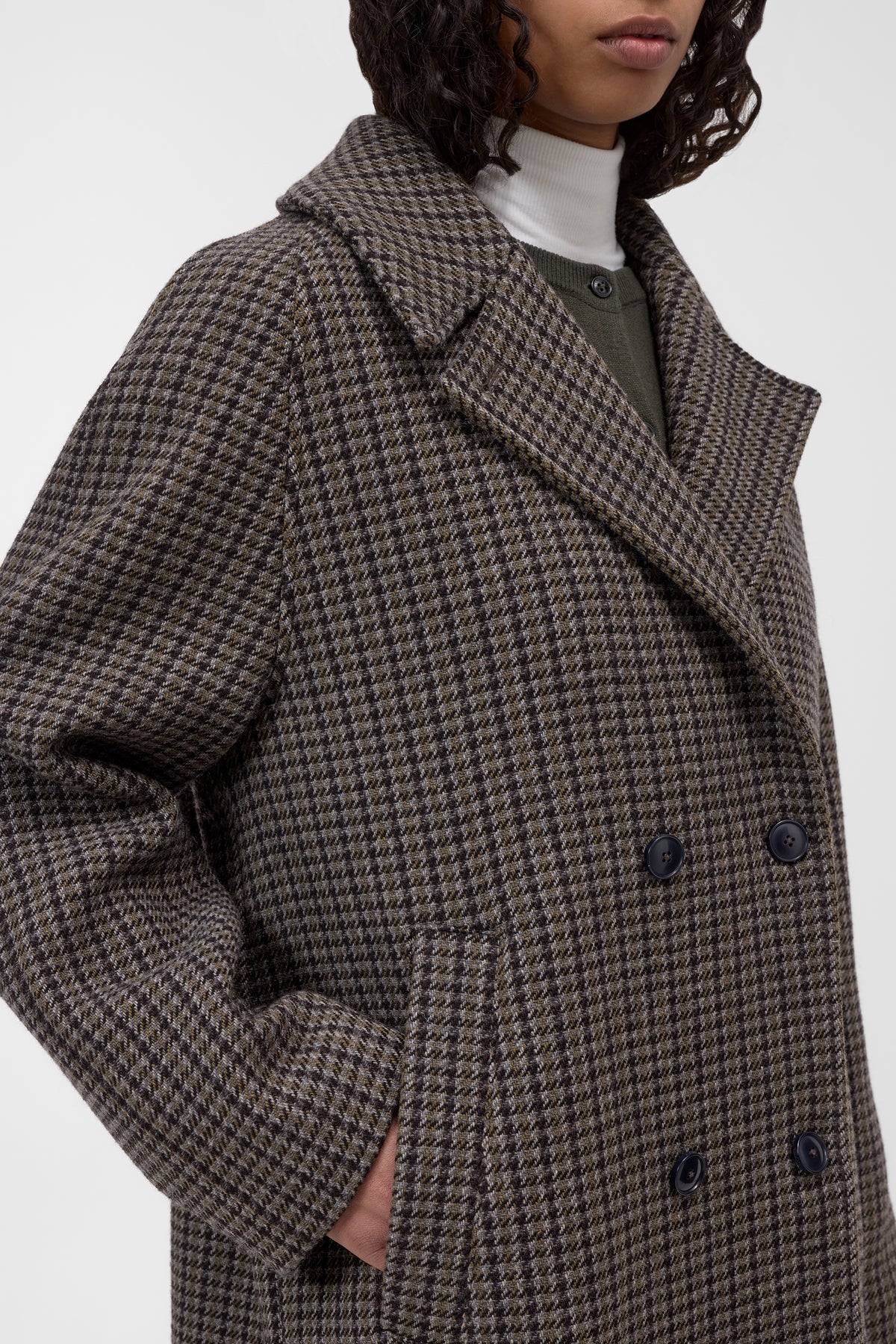 A close-up of a model wearing a double-breasted houndstooth coat in brown and beige, layered over a white turtleneck and olive green top.