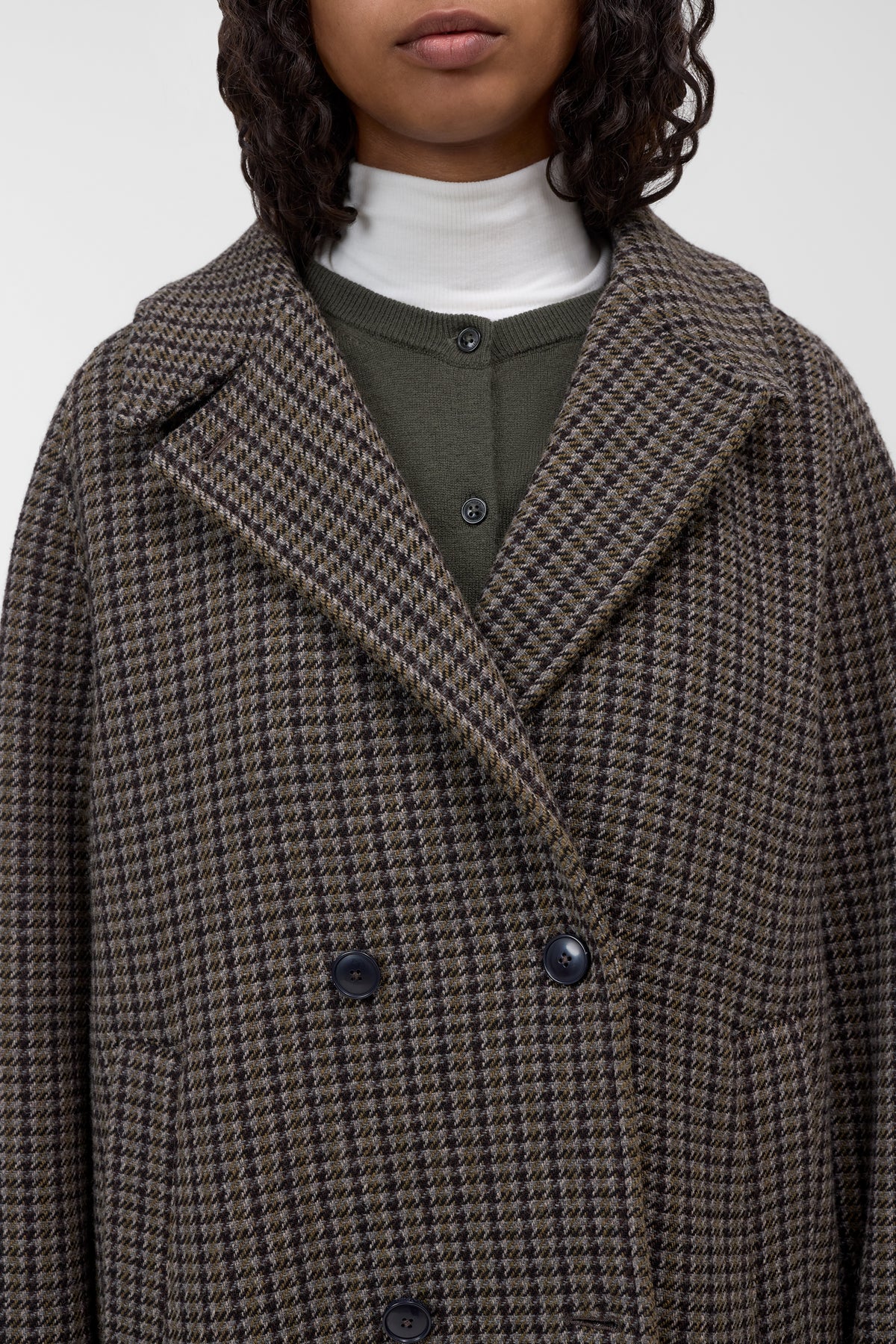 A close-up of a model wearing a brown and black houndstooth double-breasted coat over an olive green top and white turtleneck.