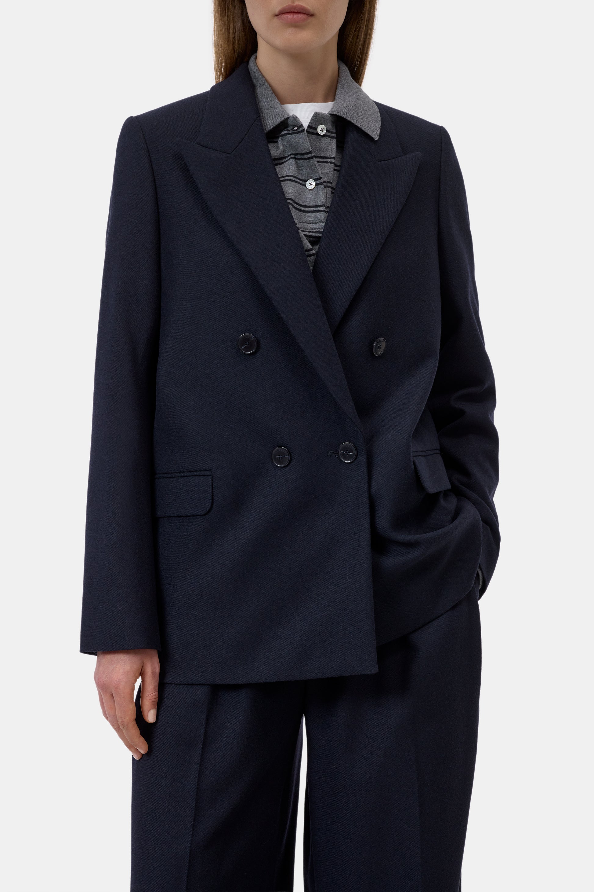 A model wearing a dark navy double-breasted blazer with matching trousers, styled with a striped shirt underneath, against a light gray studio background.
