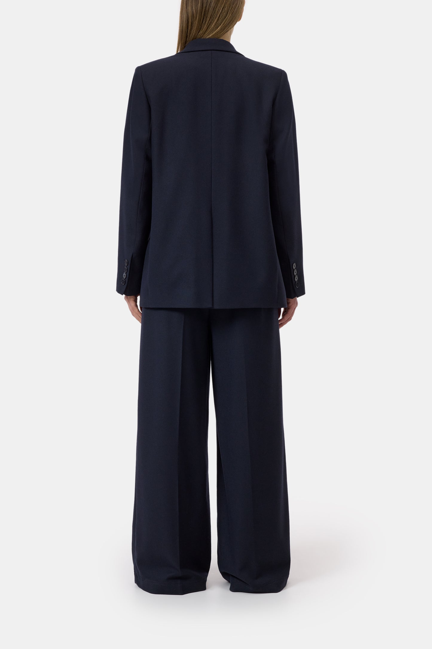 A model shown from the back wearing a dark navy blue tailored suit, featuring a structured blazer and matching wide-leg trousers, against a plain light gray studio background.