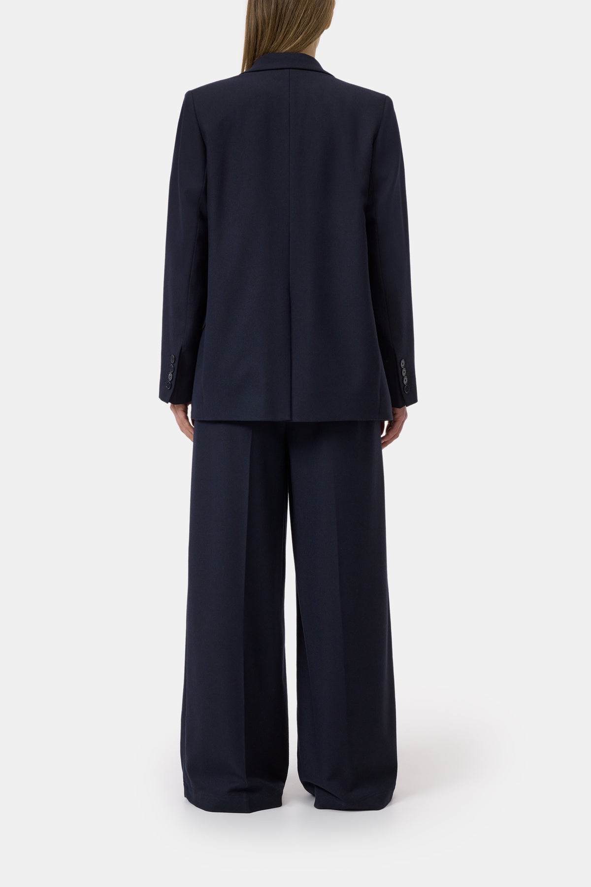 A model shown from the back wearing a dark navy blue tailored suit, featuring a structured blazer and matching wide-leg trousers, against a plain light gray studio background.