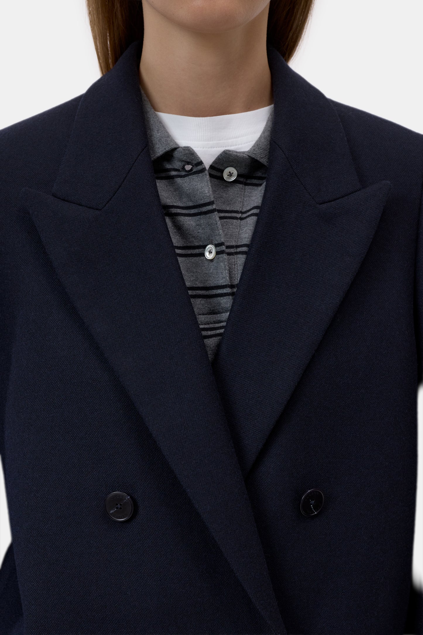 Close-up of a person wearing a dark navy double-breasted coat over a gray striped button-up shirt and a white undershirt, against a plain light background.