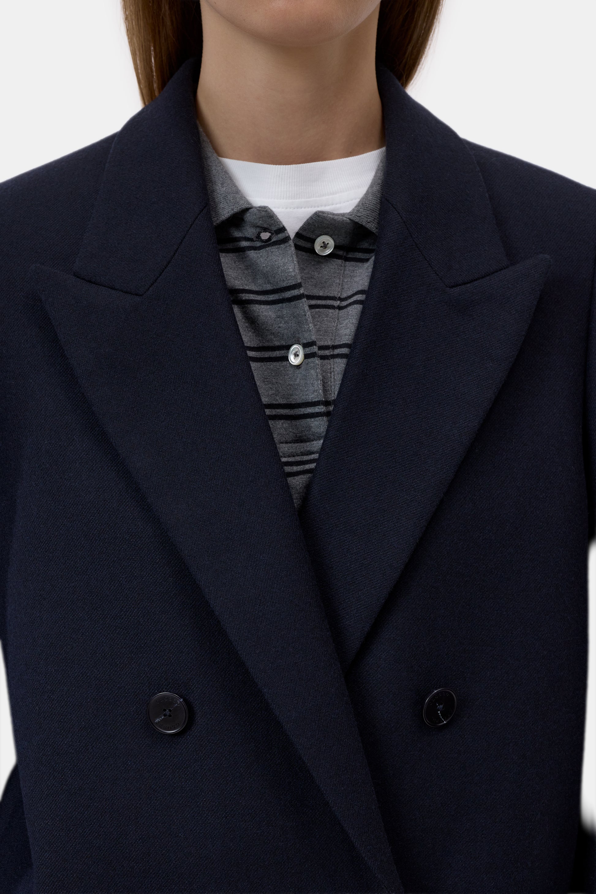 Close-up of a person wearing a dark navy double-breasted coat over a gray striped button-up shirt and a white undershirt, against a plain light background.
