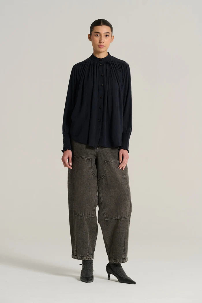 A woman shown in front profile wearing a dark navy long-sleeve blouse with gathered cuffs and grey jeans, photographed against a plain light gray studio background.