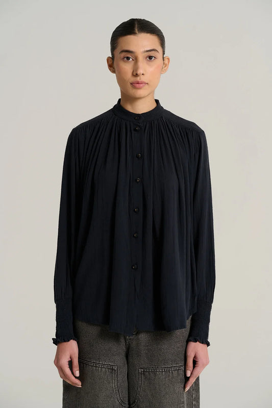 A woman shown in front profile wearing a dark navy long-sleeve blouse with gathered cuffs and grey jeans, photographed against a plain light gray studio background.