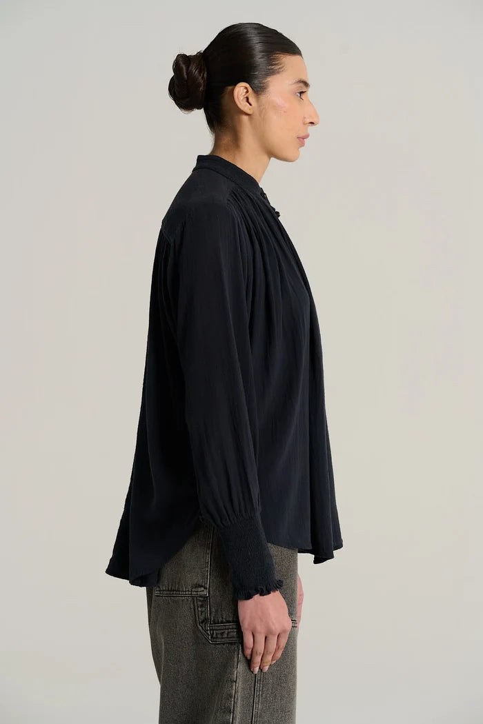 A woman shown in side profile wearing a dark navy long-sleeve blouse with gathered cuffs and grey jeans, photographed against a plain light gray studio background.