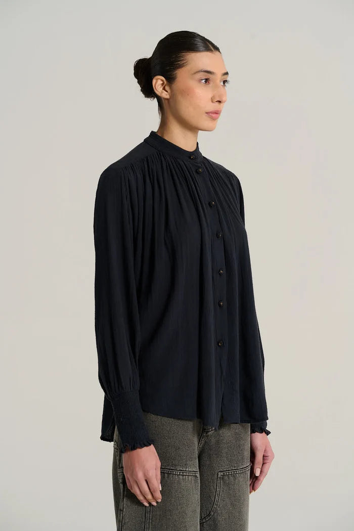 A woman wearing a dark navy long-sleeve blouse with a high gathered collar and front buttons, paired with gray pants, photographed against a plain light gray studio background.