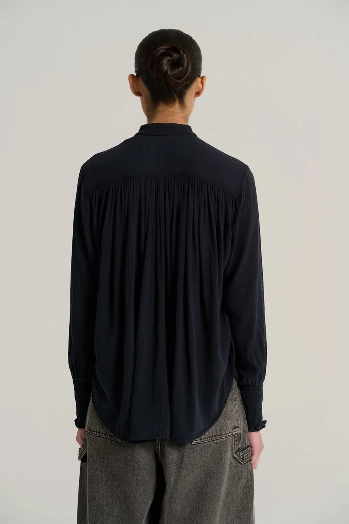 A woman shown in back wearing a dark navy long-sleeve blouse with gathered cuffs and grey jeans, photographed against a plain light gray studio background.