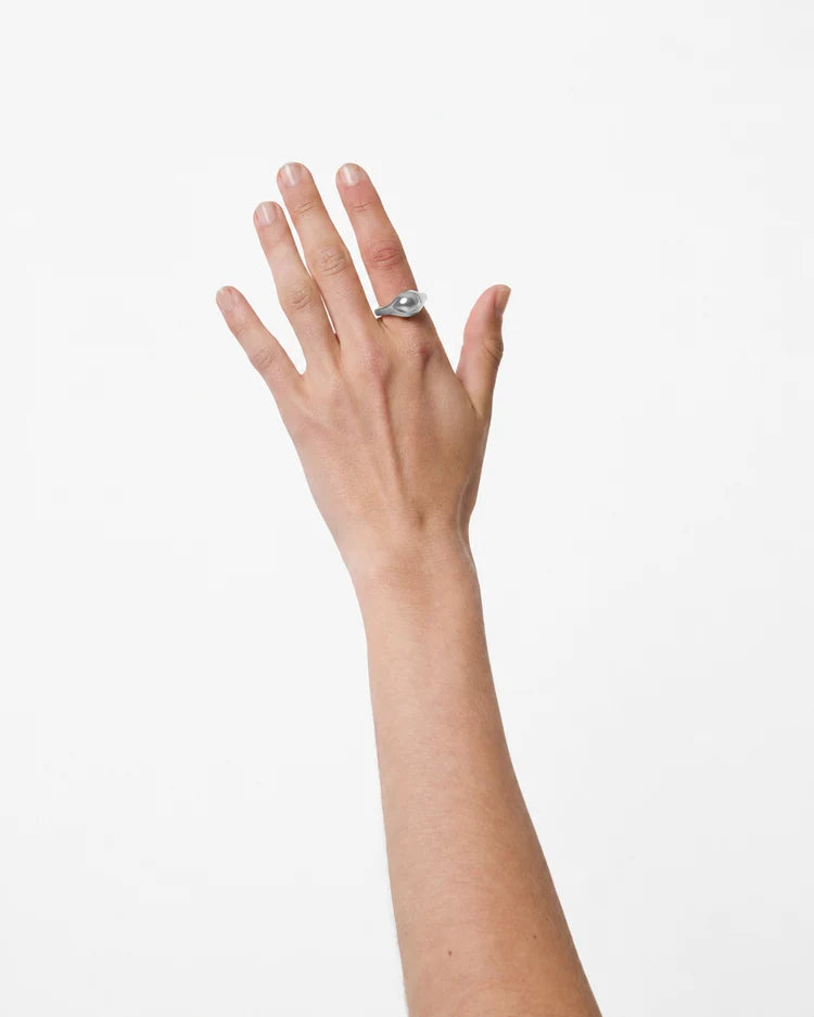 A silver ring with a curved, wave-like band and a central clear gemstone, shown on a person's index finger against a plain white background.